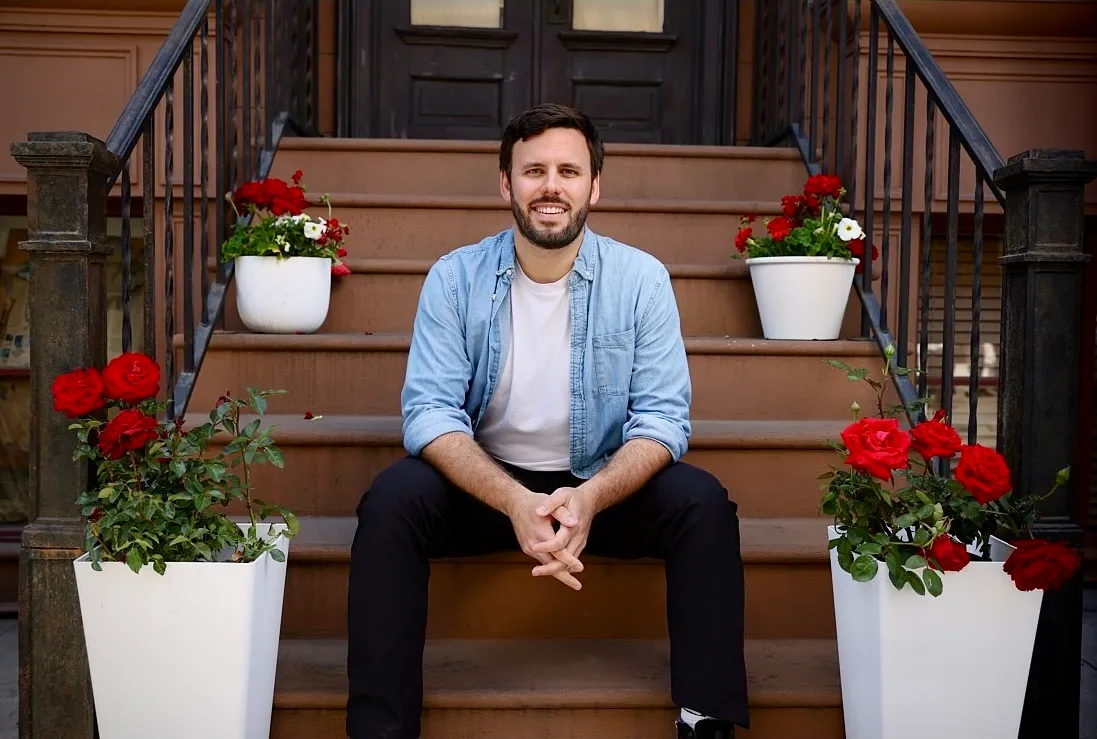 Matt Reichard sitting on a brownstone stoop between red and white flowers, wearing a light denim shirt over a white tee.